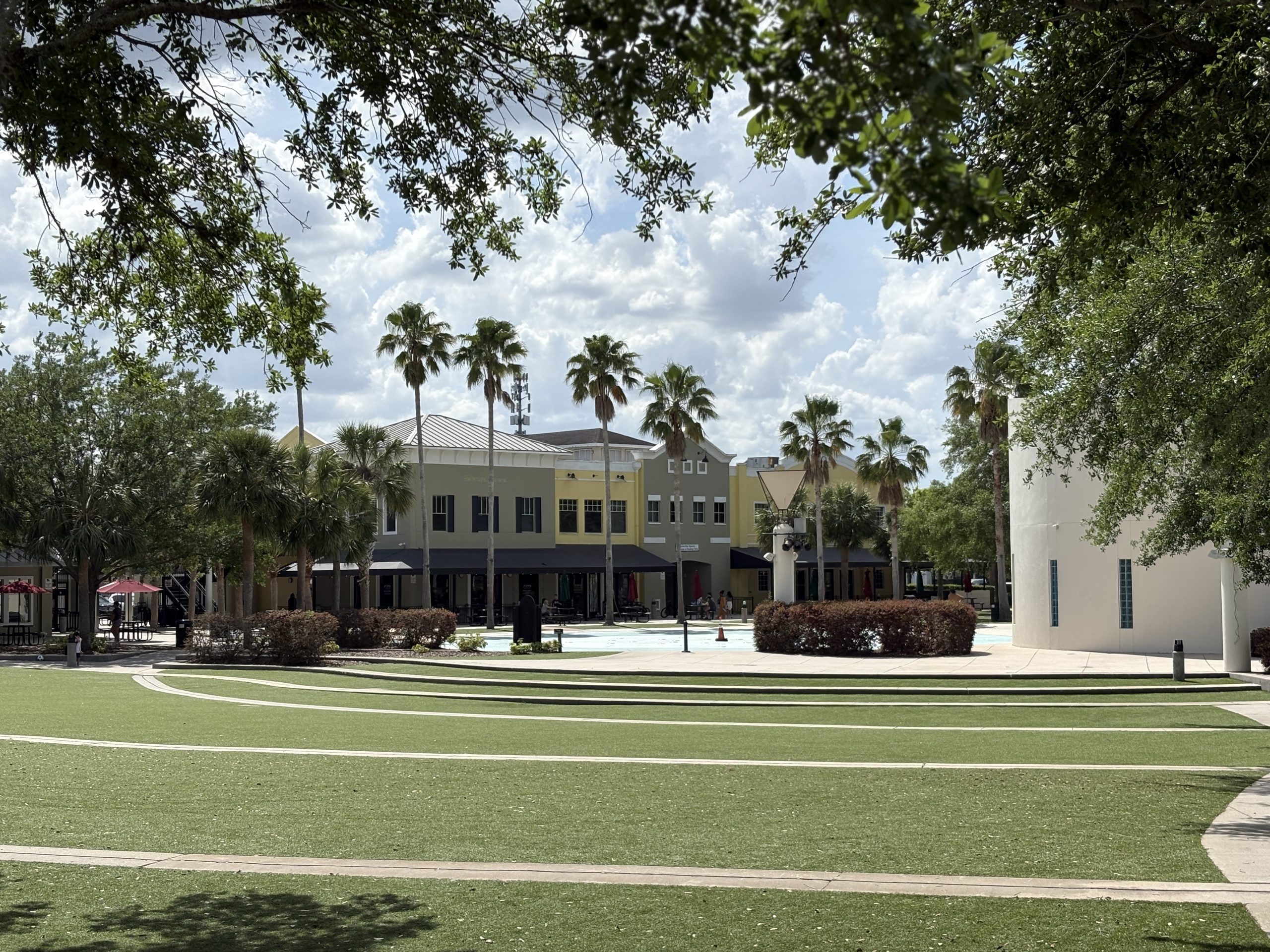 View of Park Square in the Garden District of FishHawk Ranch, featuring a grassy open area with curved pathways in the foreground, surrounded by palm trees, colorful storefronts, and shaded seating areas under a partly cloudy sky.