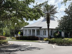 A large, light-colored building i FishHawk Ranch's Garden District with an arched entrance and the sign "The Aquatic Club" displayed above it. The building is surrounded by tall palm trees, manicured bushes, and other greenery under a partly cloudy blue sky. The scene appears to be taken in a warm, tropical location.