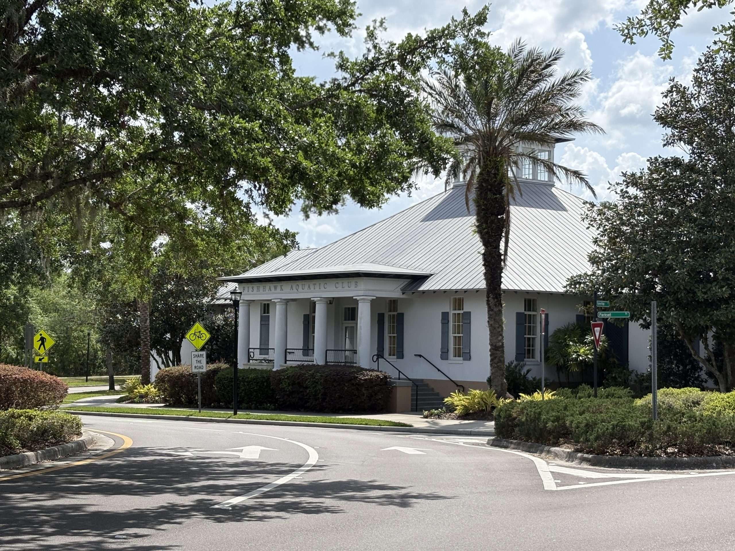 A large, light-colored building i FishHawk Ranch's Garden District with an arched entrance and the sign "The Aquatic Club" displayed above it. The building is surrounded by tall palm trees, manicured bushes, and other greenery under a partly cloudy blue sky. The scene appears to be taken in a warm, tropical location.